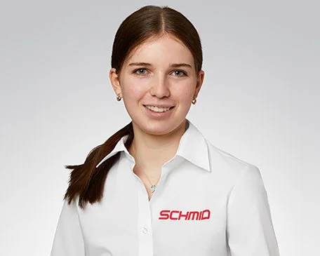 Eine junge Frau mit roten Haaren vor grünen Blättern, trägt ein weißes Hemd, lächelt leicht.