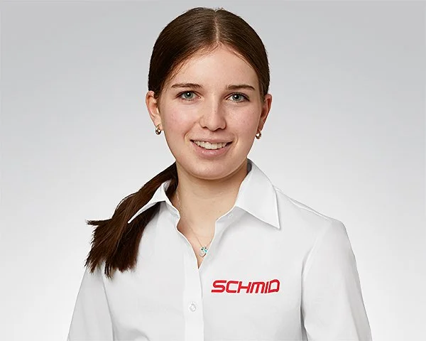Eine junge Frau mit roten Haaren vor grünen Blättern, trägt ein weißes Hemd, lächelt leicht.