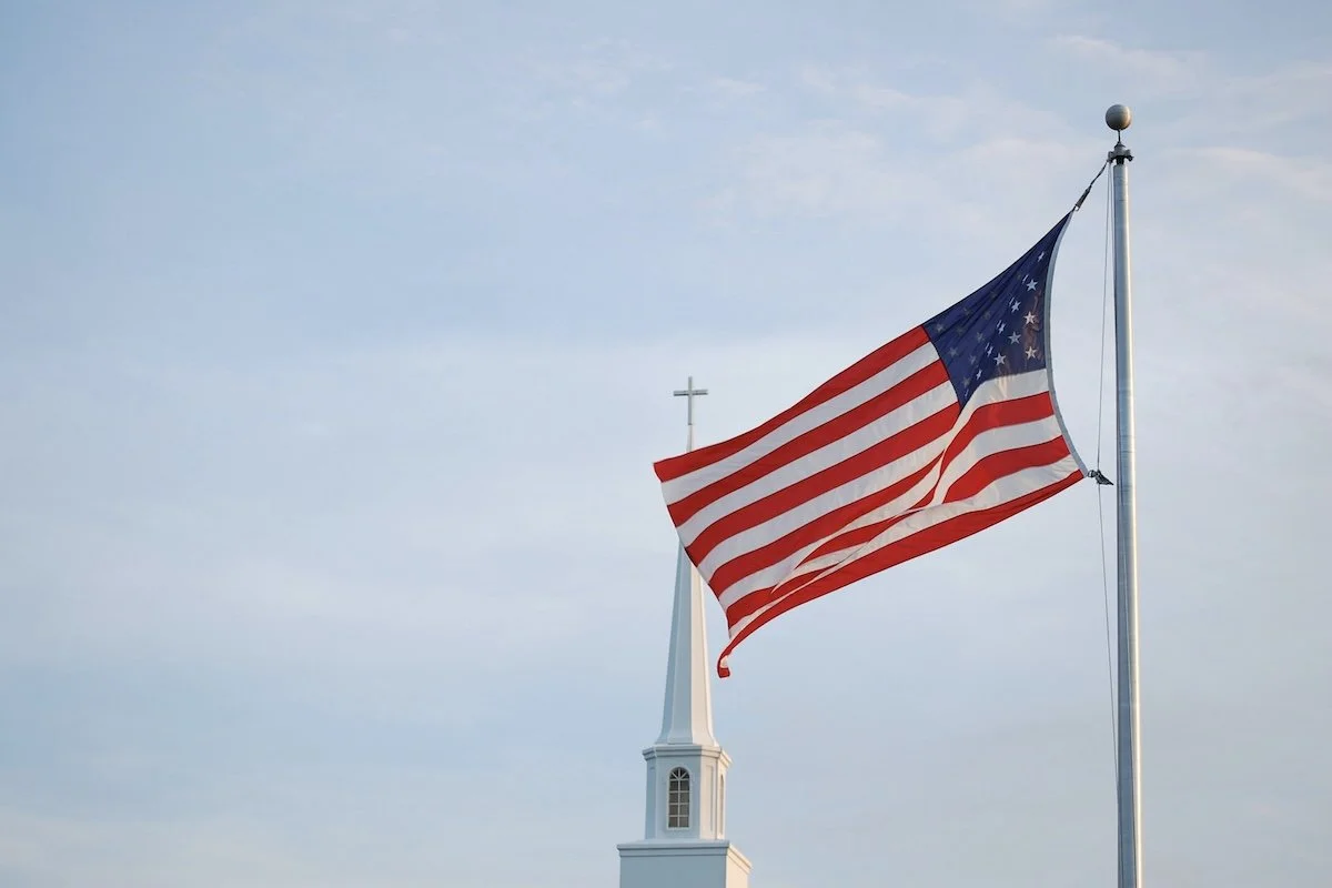 American flag waving with church steeple in the background symbolizing politics overtaking religion