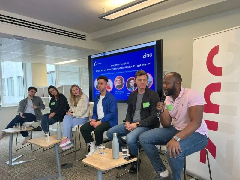 A diverse group of six young adults participating in a panel discussion in a modern conference room. All are seated on chairs, with small tables in front holding water bottles and glasses. The background features a large screen displaying investment insights related to zinc, and a vertical banner with red text. One man on the right is speaking into a microphone.