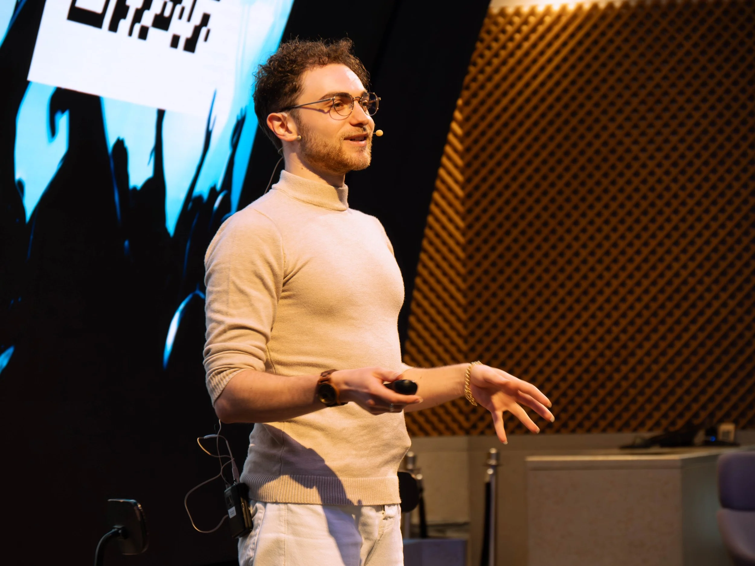 Seven Jacobs giving a presentation on stage, holding a remote control, with LED display behind him. He is wearing glasses, a beige turtleneck sweater, and beige pants.