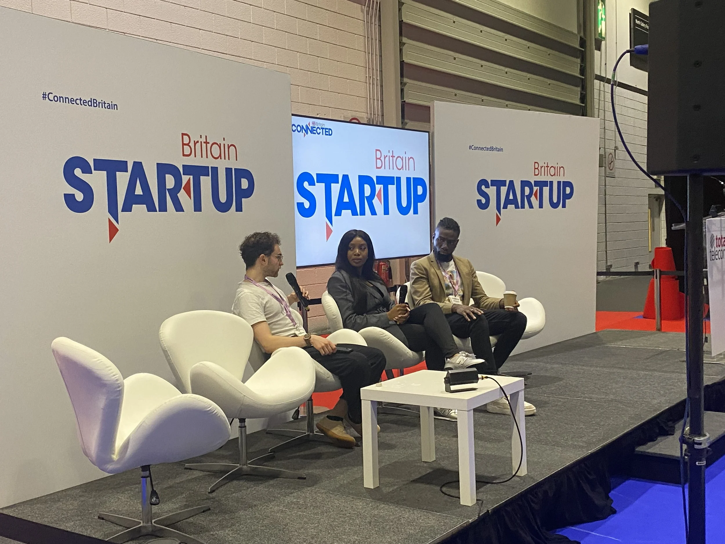 Three people sitting on a stage with a backdrop reading 'Britain Startup' and '@ConnectedBritain' hashtag. Two men and a woman, engaged in conversation with microphones and a coffee cup, seated in modern white chairs.