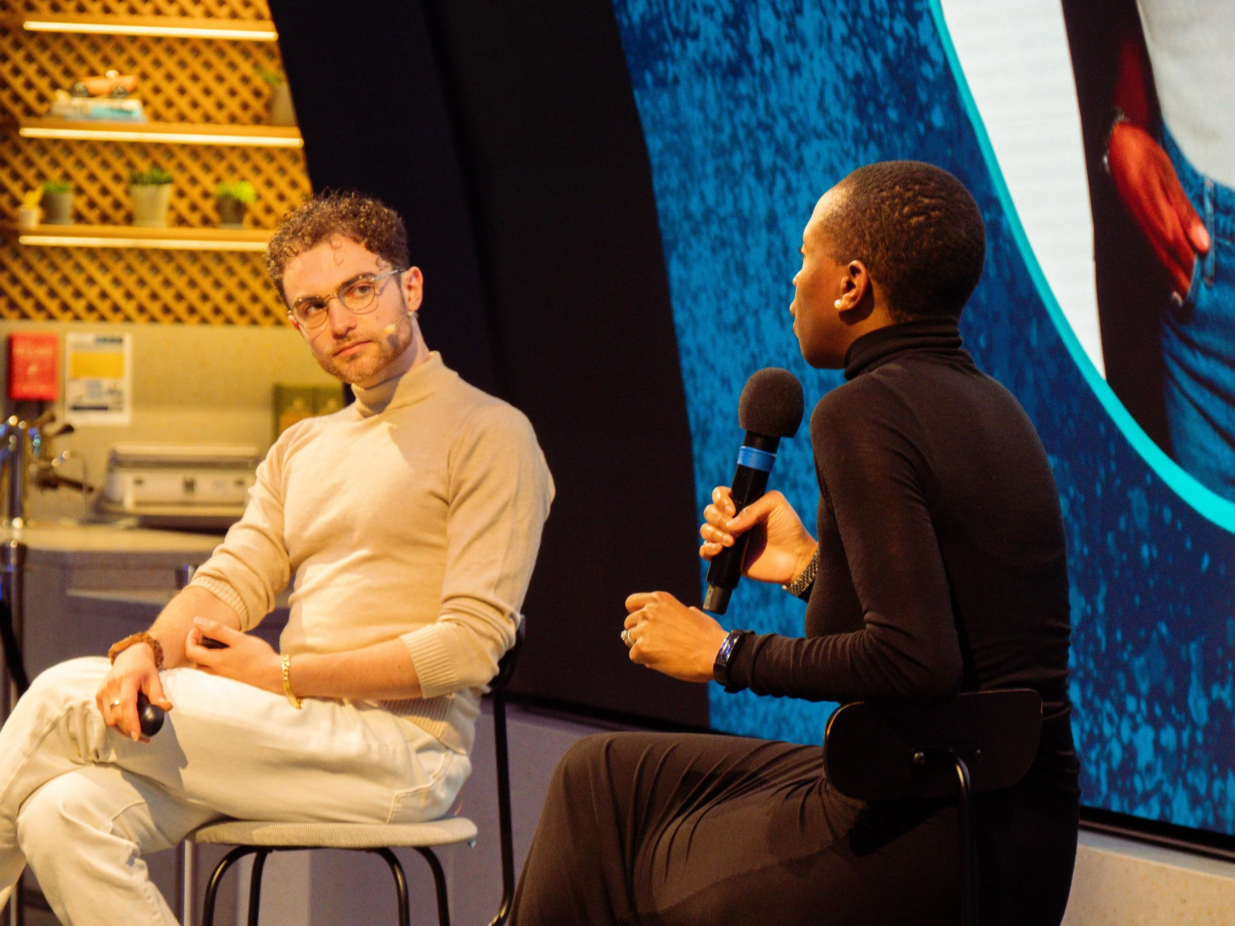 Seven Jacobs seated on stage engaged in a discussion, with one person holding a microphone. The background features a large screen and shelves with small potted plants. The setting appears to be a conference or talk event.