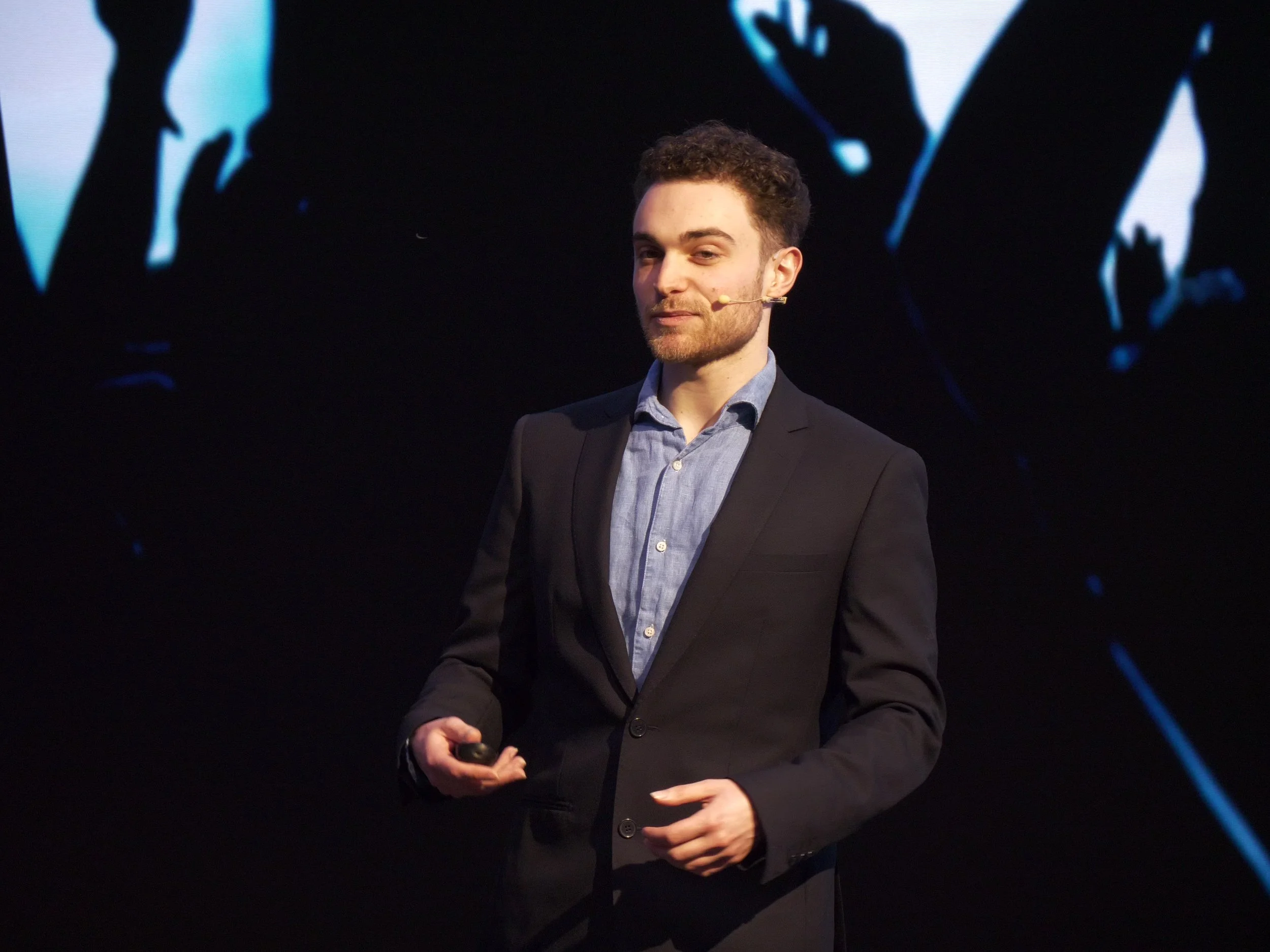 Seven Jacobs in a suit giving a presentation on stage, holding a clicker in his right hand, with a dark background and blue abstract designs.