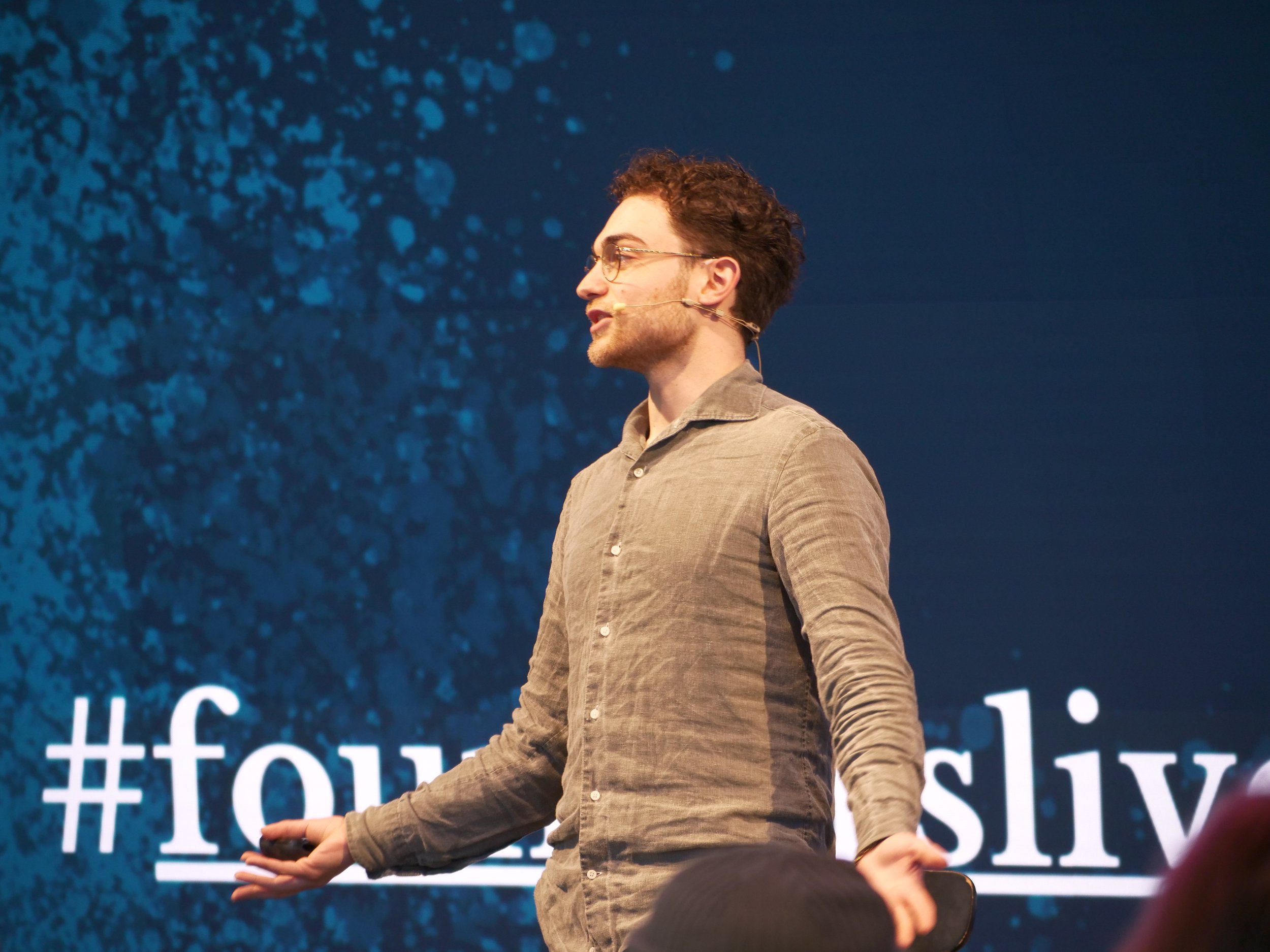 Seven Jacobs speaking on stage with a blue background and white text that reads '#foursl'.