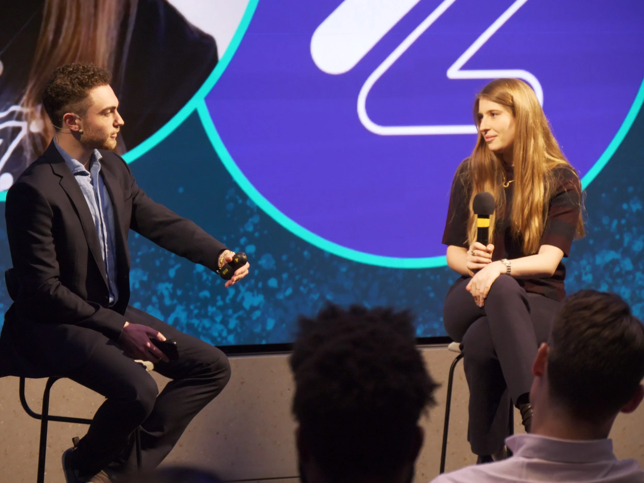  in a dark suit is sitting on a stool on a stage, holding a microphone, engaged in conversation with a young woman in a black outfit, who is also sitting on a stool and holding a microphone. They are in front of a large screen with a blue and purple 