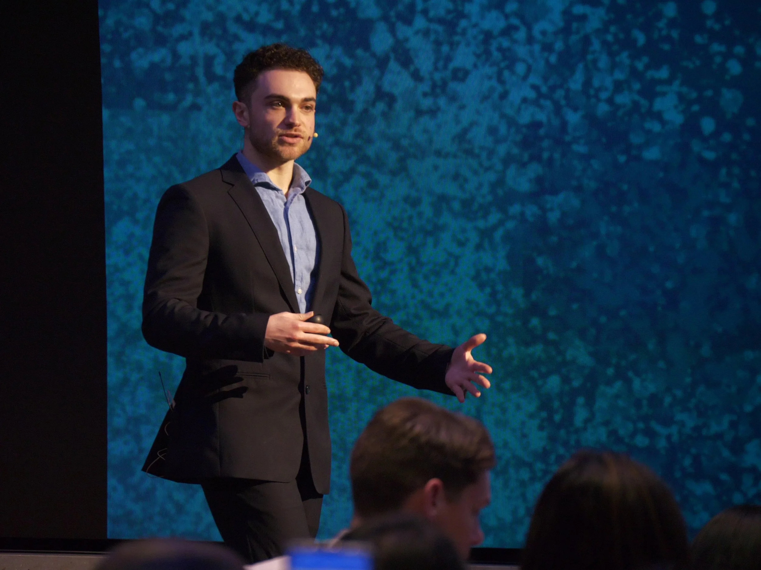 A man in a dark suit presenting on stage with a blue textured background.