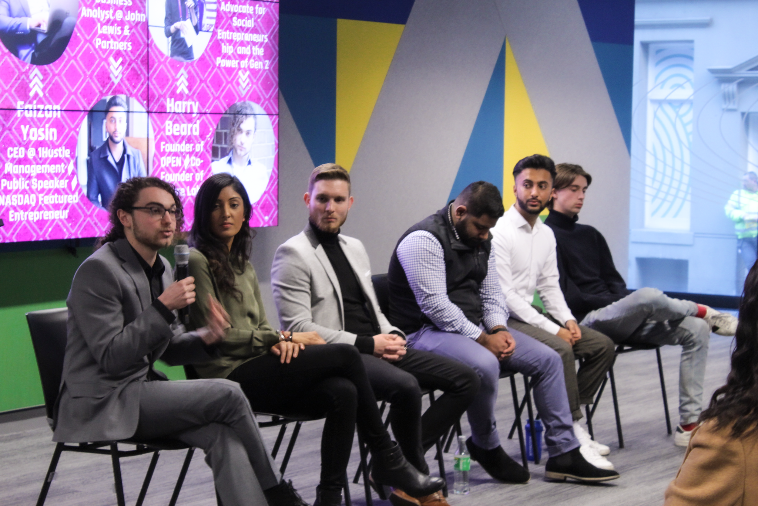 Seven Jacobs one of six people seated on chairs at a panel discussion, with a pink and colorful background displaying headshots and bios of speakers, including titles and names.