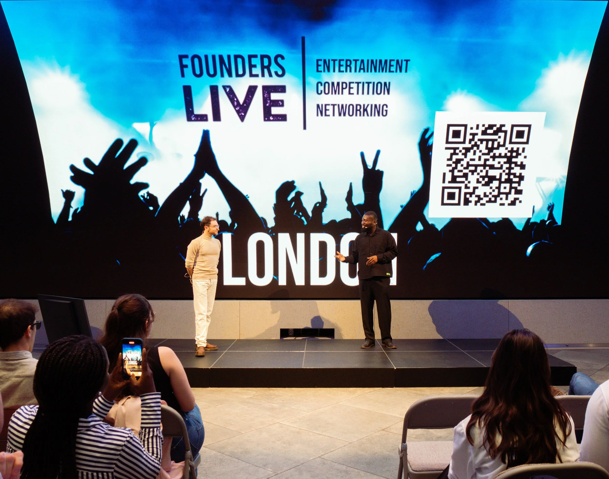 Two presenters on stage at a conference in front of a large screen with a crowd watching, the screen displays text about founders live, entertainment, competition, networking, a QR code, and an audience in a conference room.