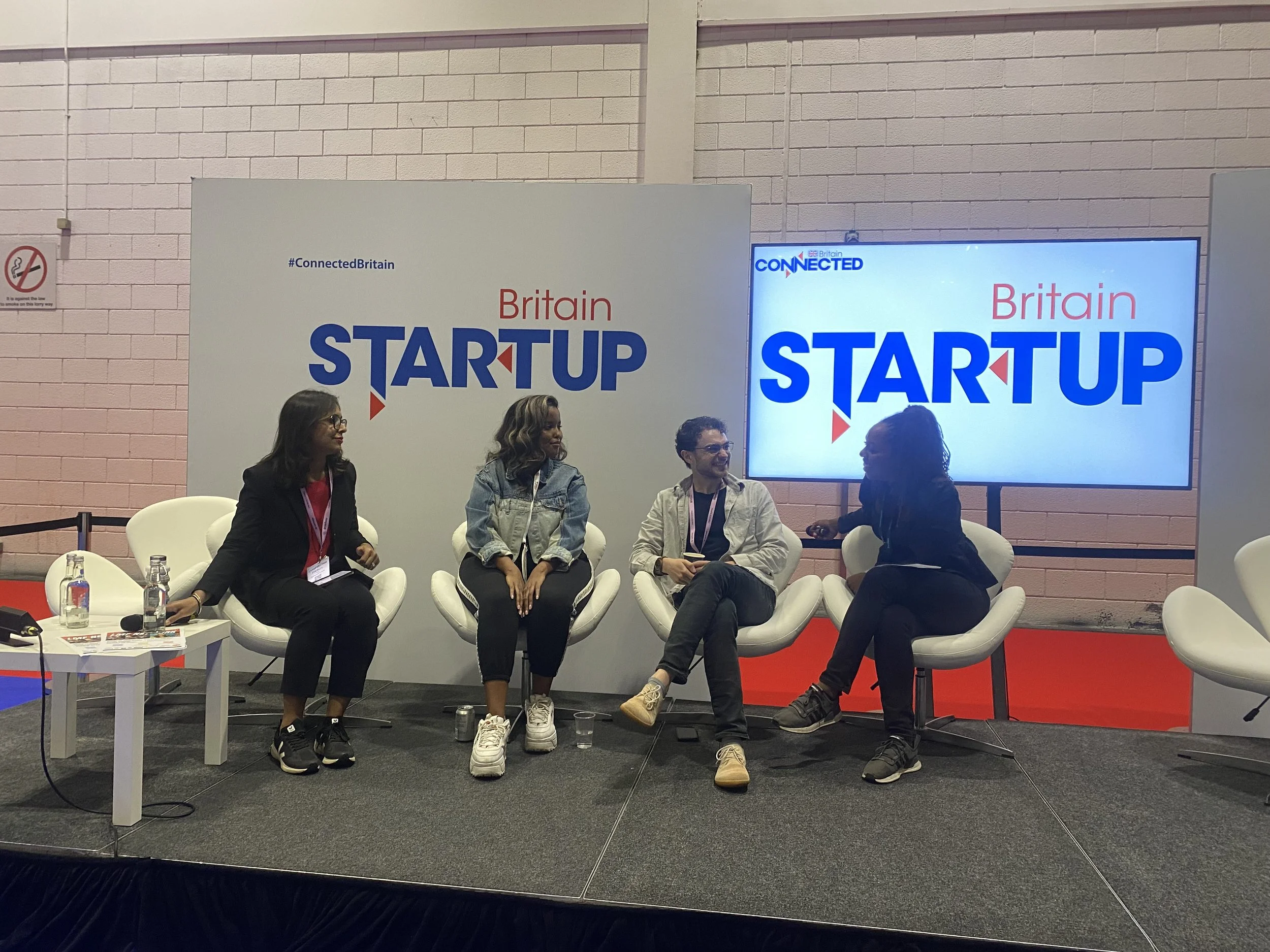 Four people sitting on a stage at a startup conference, engaging in a discussion. Behind them, a large screen displays 'Britain STARTUP' and the hashtag #ConnectedBritain.