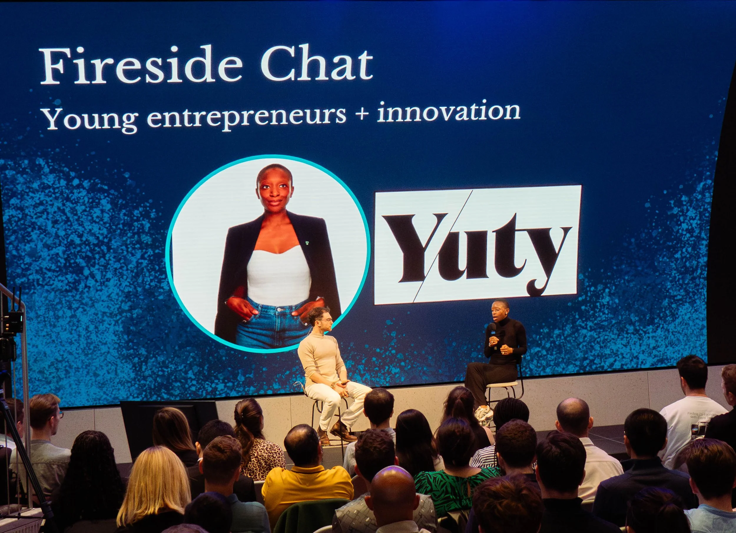 A large screen shows the title 'Fireside Chat' with the subtitle 'Young entrepreneurs + innovation.' There is a photo of a woman with a short haircut, wearing a white top and black blazer, next to the Yuty logo. Two women are on stage, one with short