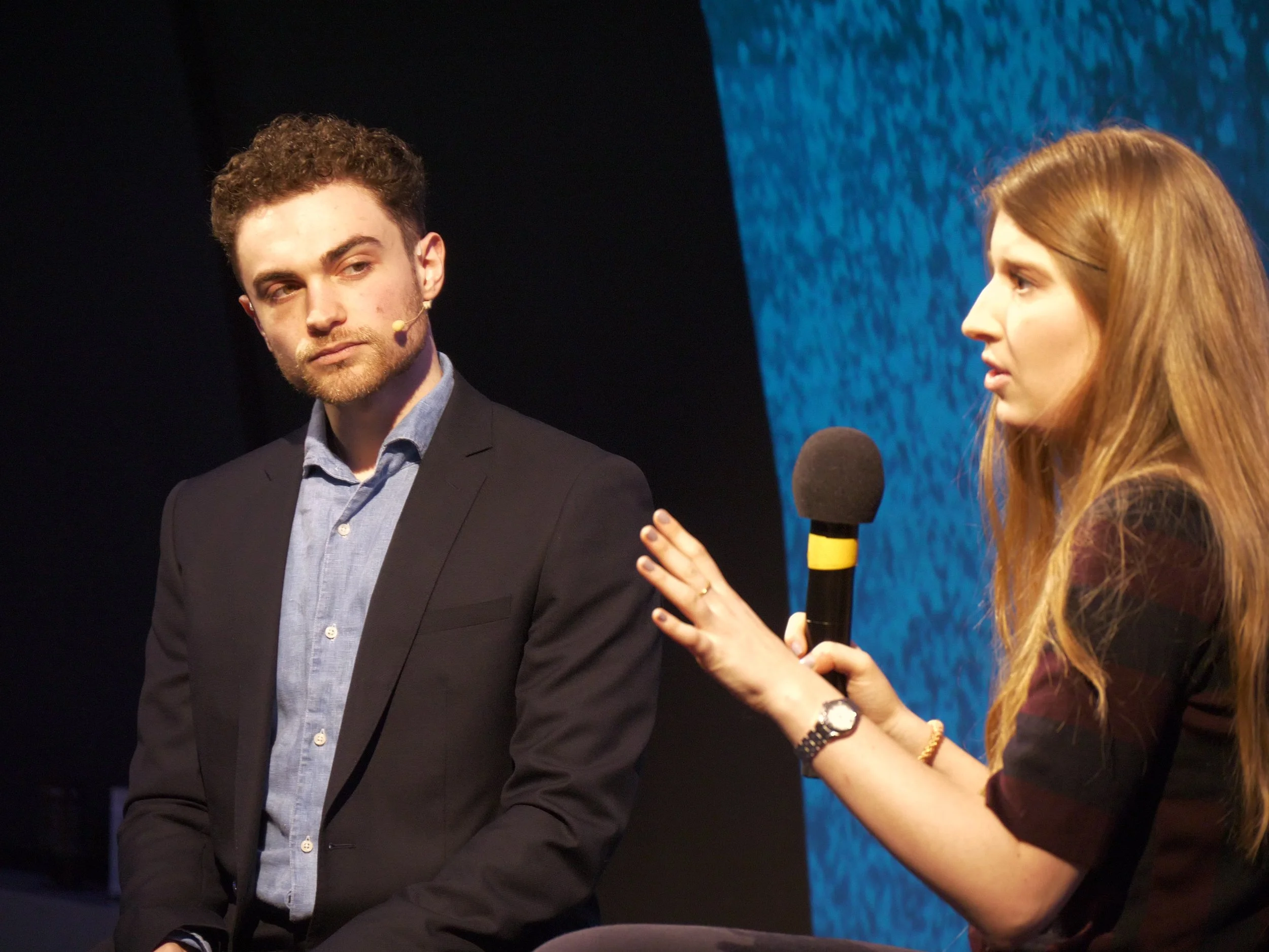 Two people on stage, one woman speaking into a microphone and Seven Jacobs listening, in a formal setting.