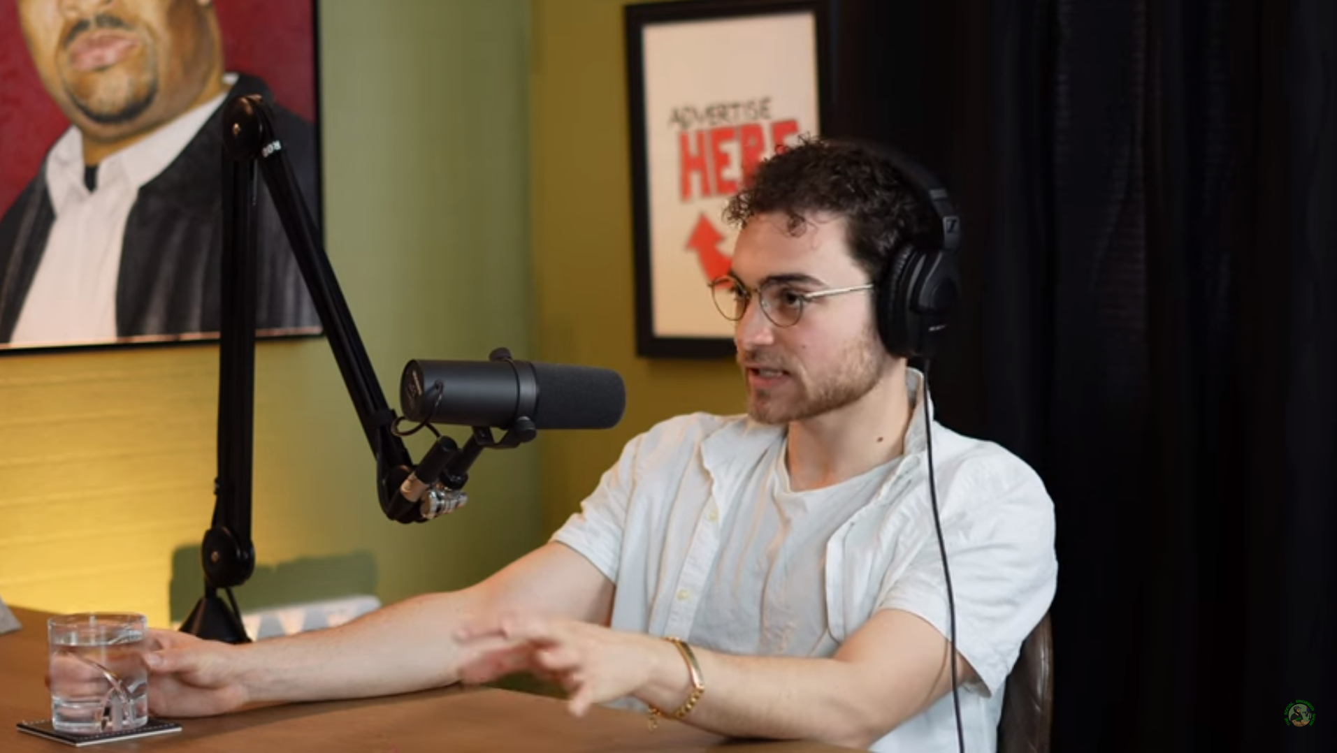 A young man with glasses and curly hair wearing a white shirt speaking into a microphone during a podcast recording. He has headphones on and is gesturing with one hand. On the wall behind him, there are framed posters, one of which says "Advertise Here" in red.
