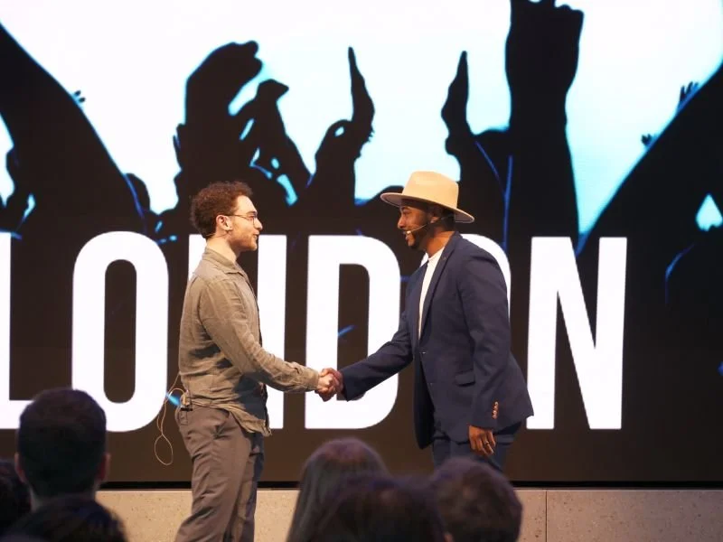 Two men shaking hands on stage in front of a large screen with silhouettes of people and the word "FOUND" in the background. One man is wearing glasses and a beige shirt, and the other is wearing a wide-brimmed hat and a blazer.