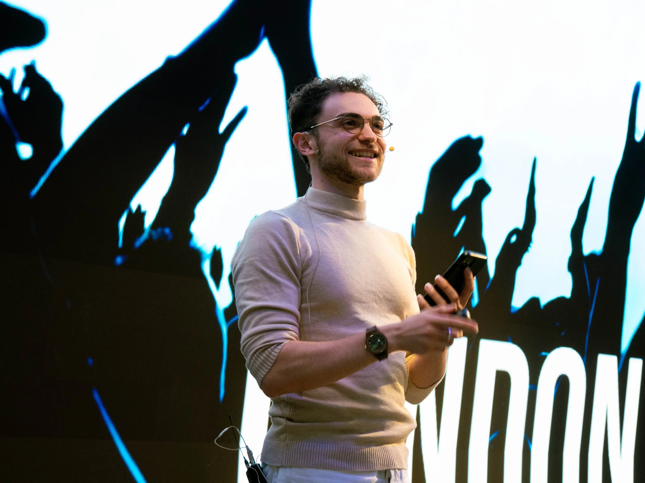Seven Jacobs smiling and holding a smartphone, standing in front of a large screen with a graphic background.