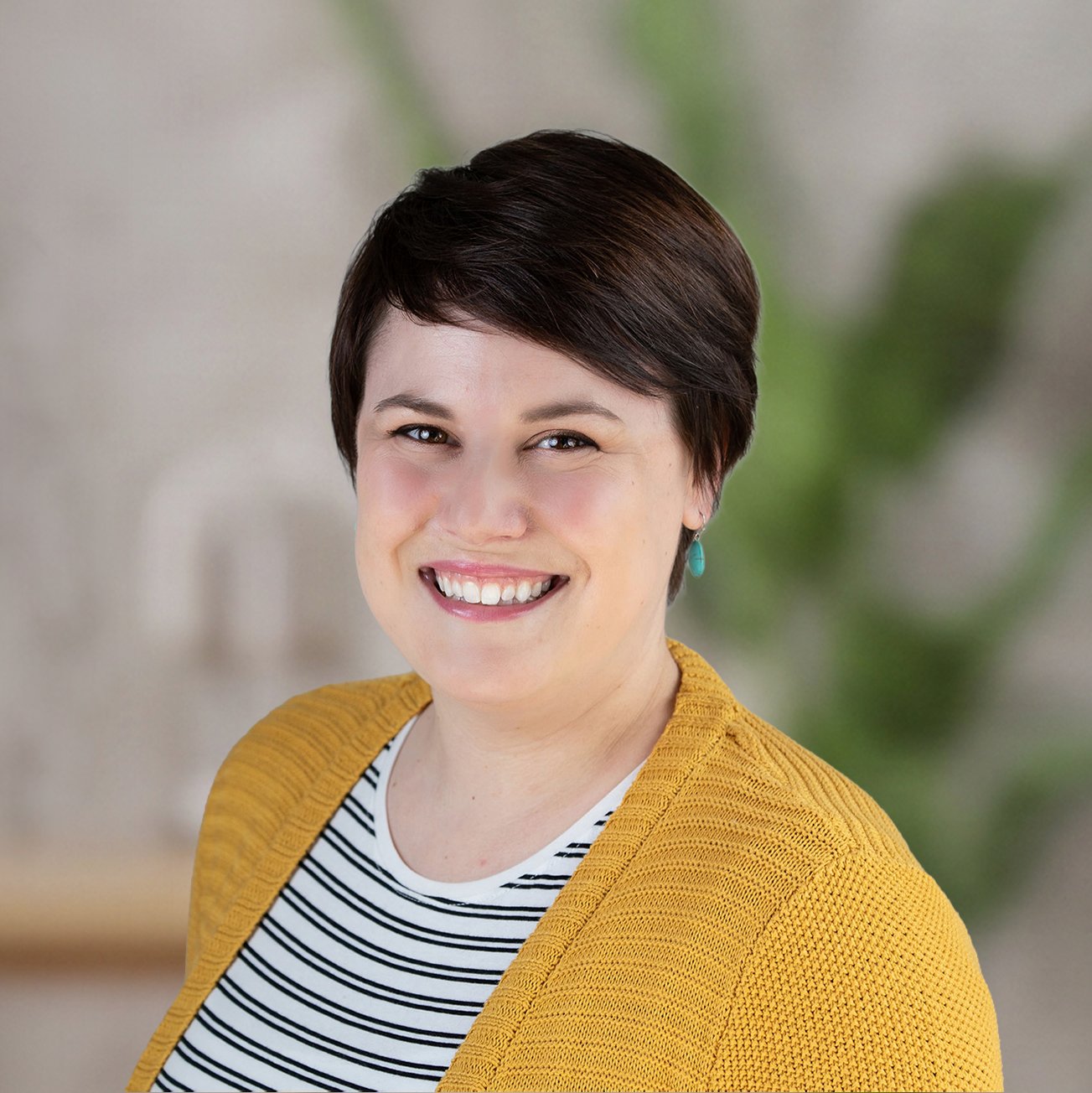 Headshot of a smiling woman with short dark hair, wearing a striped shirt, yellow cardigan, and turquoise earrings, against a blurred indoor background with green plant elements.