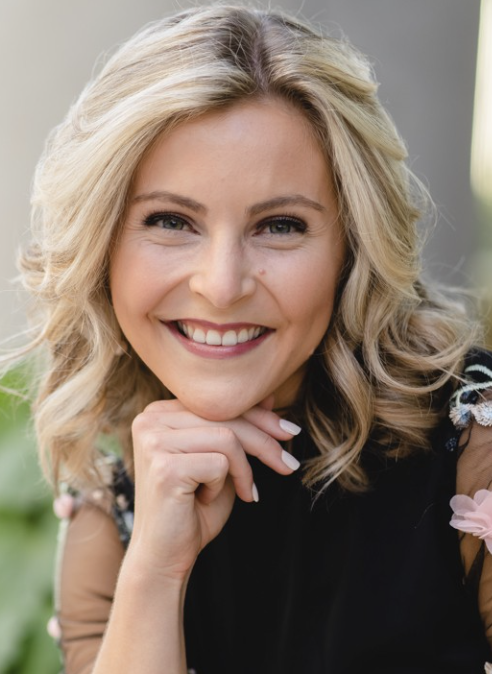 A smiling woman with blonde hair, resting her chin on her hand, wearing a black top with floral accents.