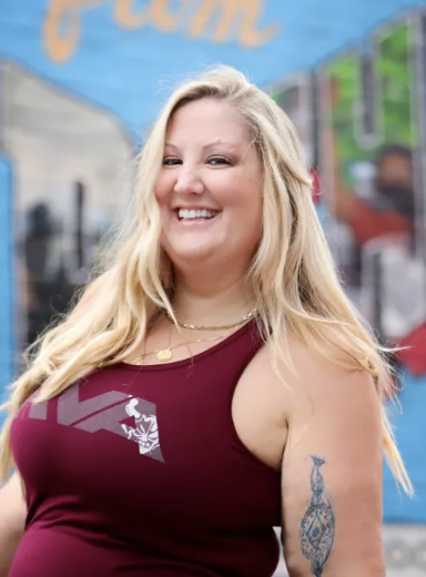 A smiling woman with long blonde hair, wearing a burgundy tank top and gold jewelry, standing outdoors in front of a colorful mural.