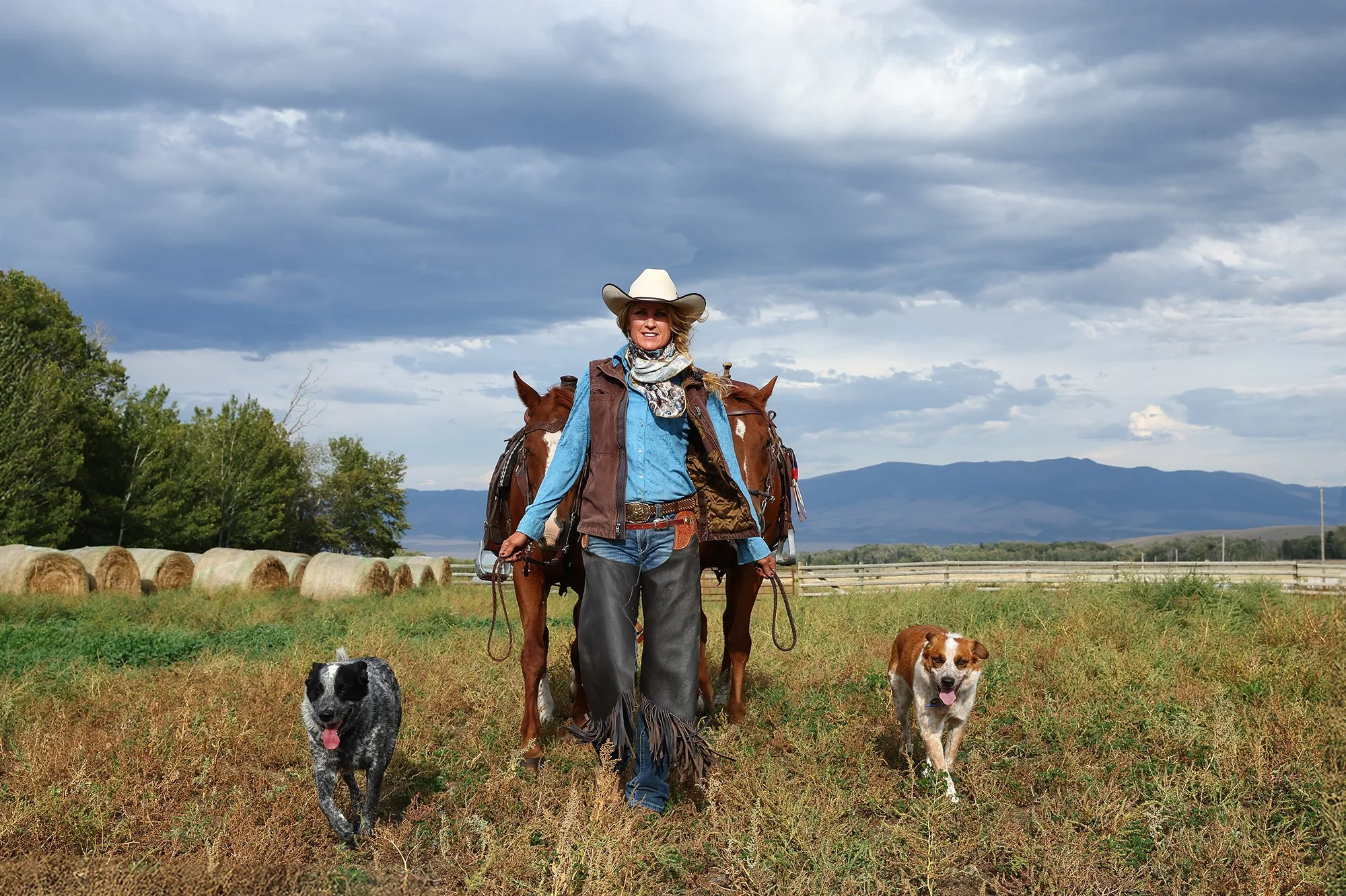Female Montana cowgirl with two horses and two dogs walking toward the camera across a field with mountains in the background under a dramatic sky