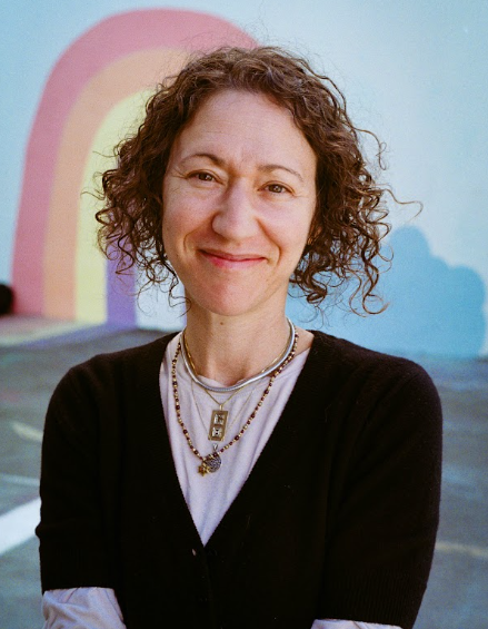 A woman with curly brown hair smiling, wearing layered necklaces and a black cardigan, standing outdoors in front of a painted rainbow mural.