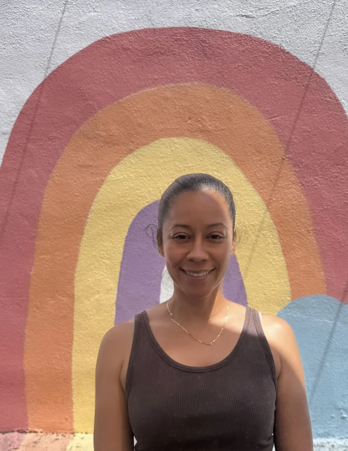 Young woman with long wavy brown hair smiling, standing in front of a painted rainbow on a wall.