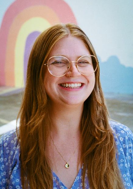 A young woman with red hair and glasses smiling in front of a colorful rainbow mural.
