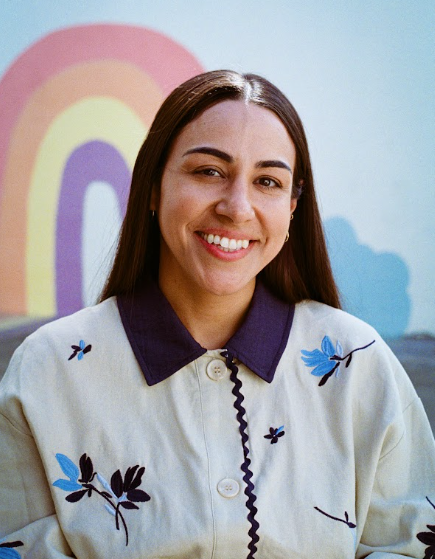A young woman with long brown hair smiling at the camera, wearing a white jacket with blue floral embroidery, standing in front of a colorful rainbow mural painted on a blue wall.