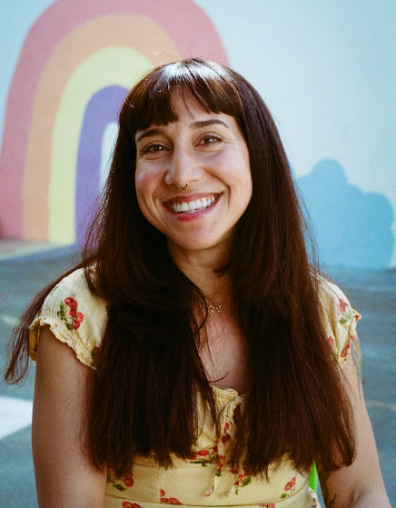 A woman with long dark hair and bangs, smiling, against a colorful mural background.