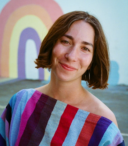 Young woman with short brown hair smiling, wearing a colorful striped shawl, standing in front of a rainbow mural.