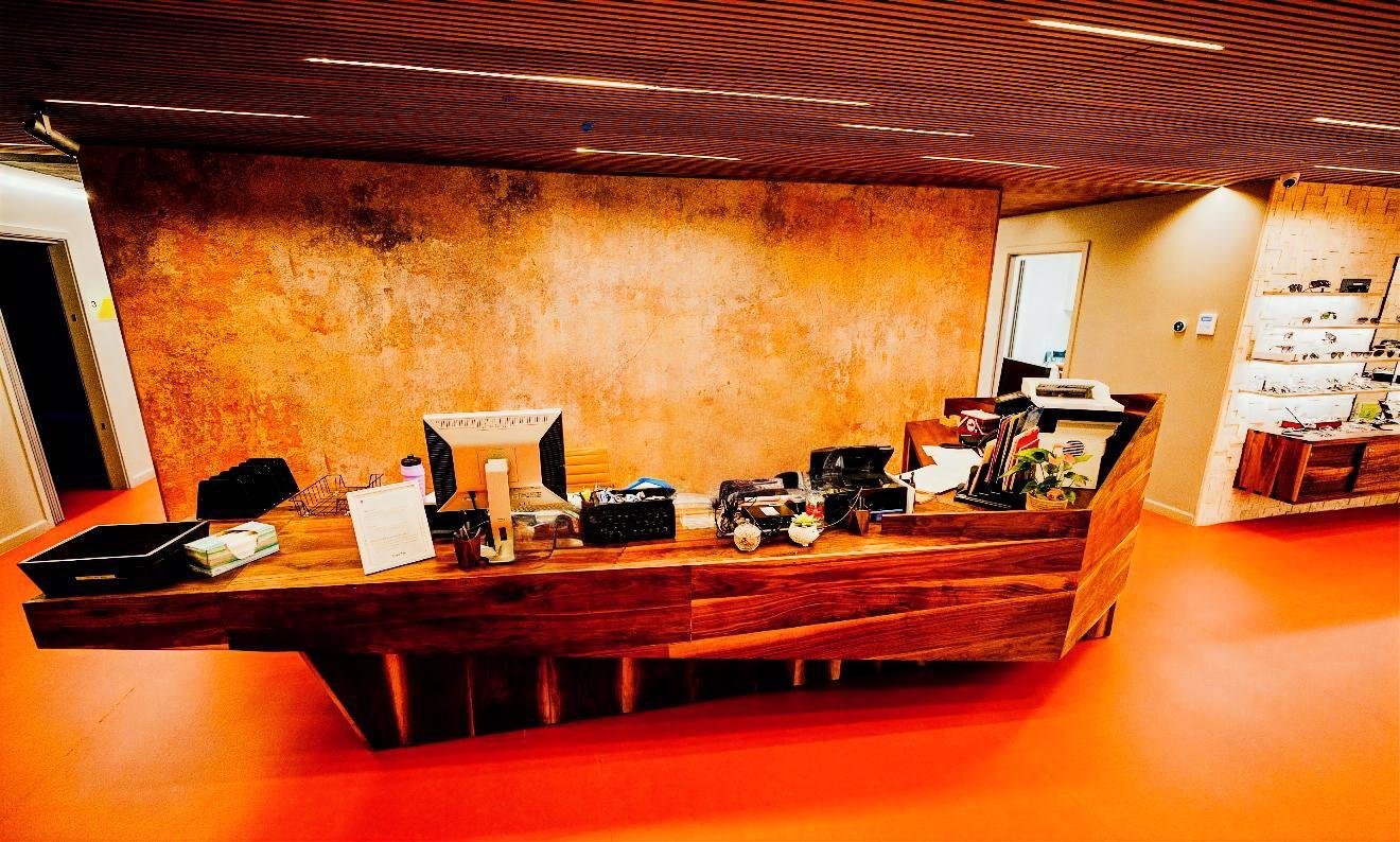 Office reception desk with various office supplies and papers on a wooden counter, a warm orange textured wall behind, and a wooden ceiling with recessed lighting.