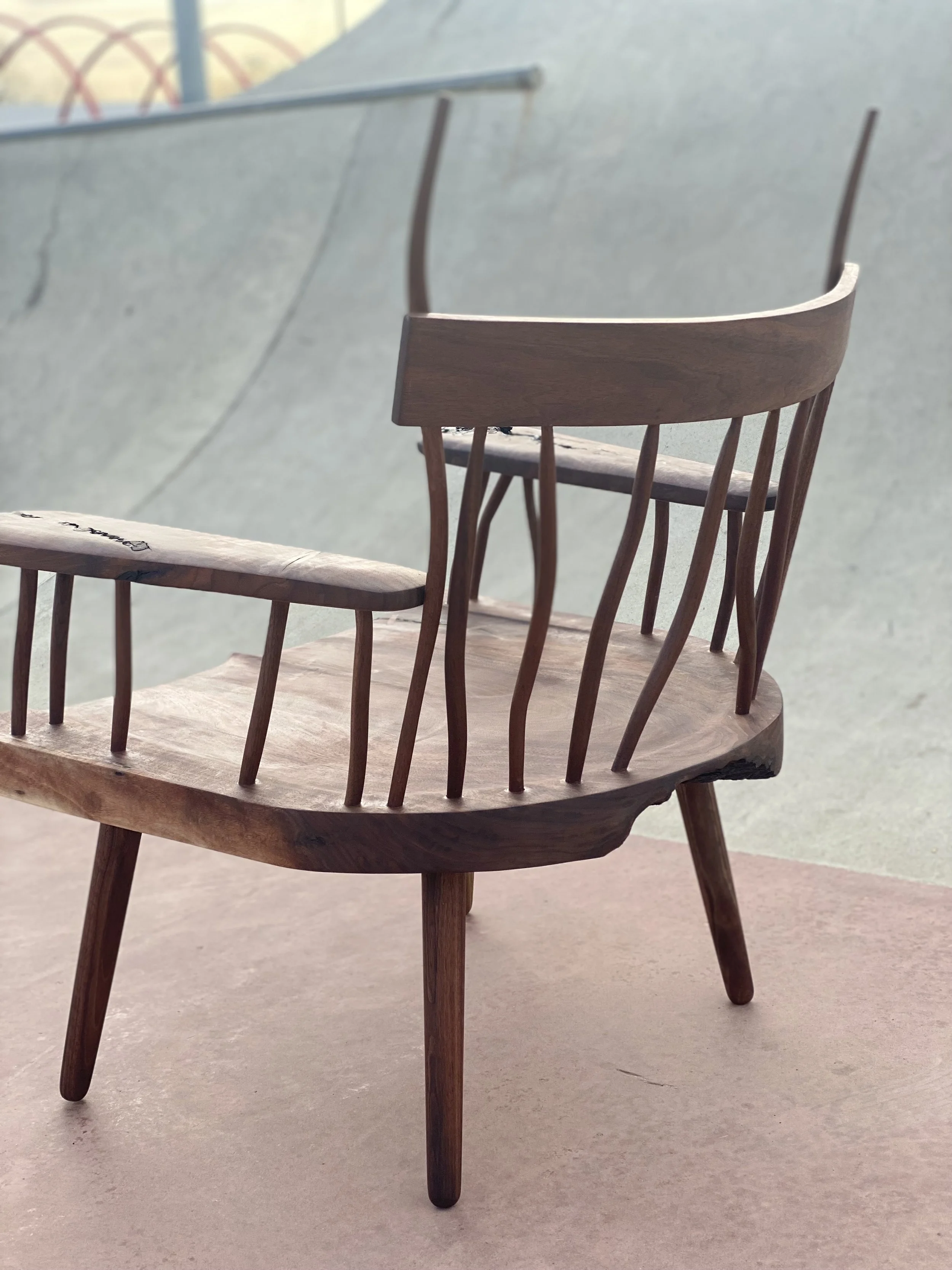 A wooden chair with a curved backrest and vertical slats, placed on a concrete surface at a skate park with a ramp in the background.
