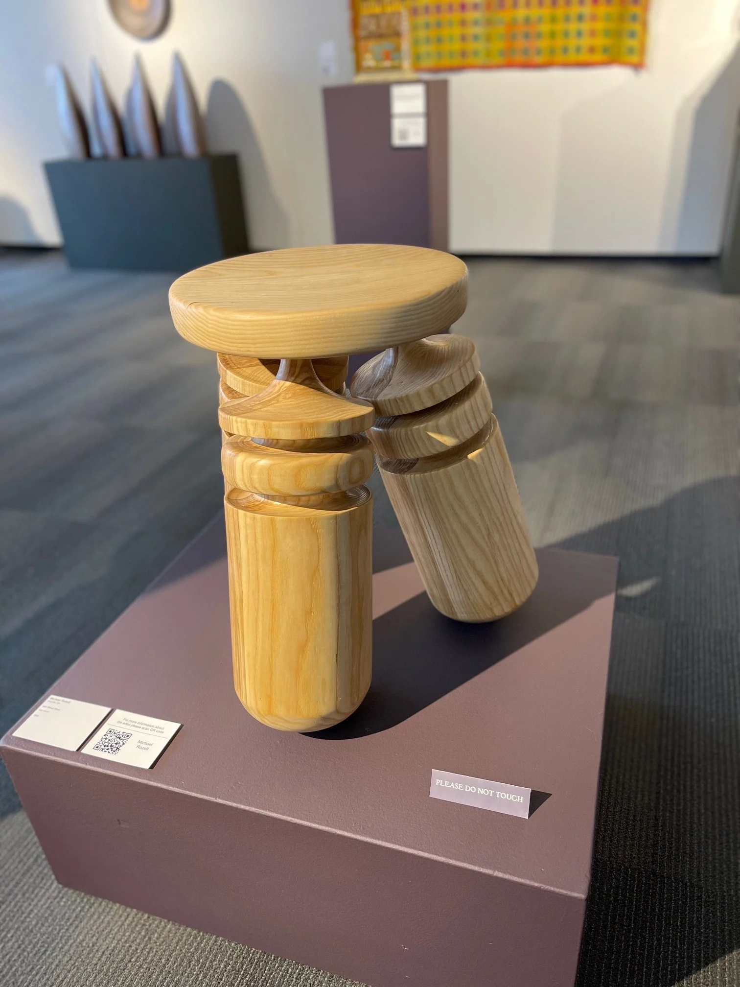A group of small wooden stools stacked on top of each other, displayed on a purple pedestal in a museum or gallery setting.