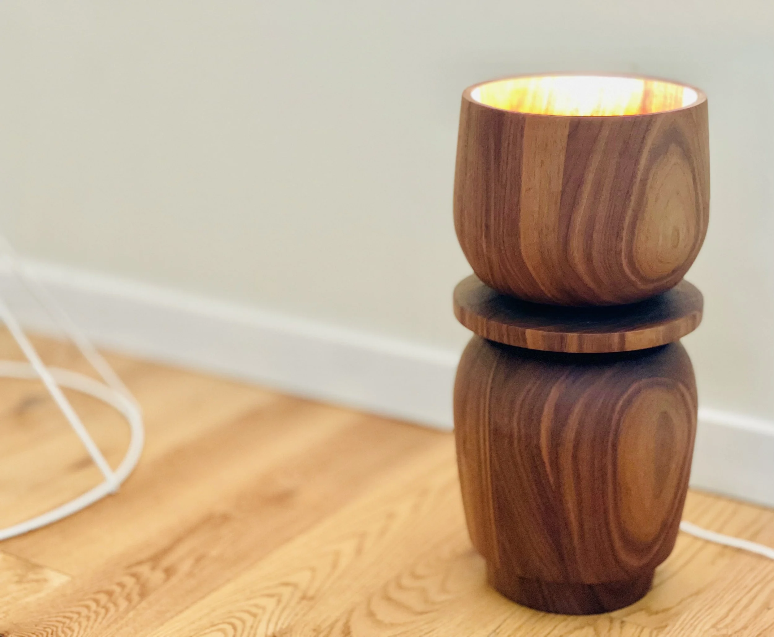 A wooden lamp with a rounded design, placed on a wooden floor against a cream-colored wall with a white baseboard.