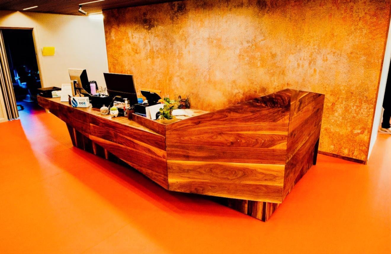 Wooden reception desk with computers, plants, and office supplies in front of a textured orange wall on an orange floor.