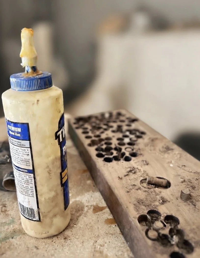 A bottle of glue placed next to a wooden plank with multiple small holes, on a dusty work surface.