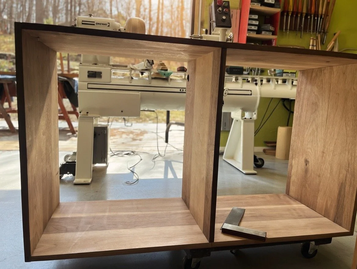 A partially assembled wooden shelf placed on a table in a workshop with woodworking tools and machinery in the background.