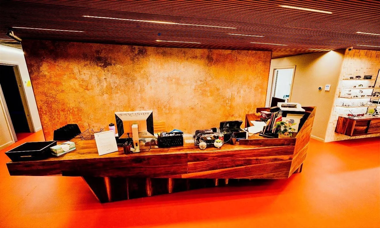 Office reception desk with various office supplies, framed photos, and plants in a modern space with a textured orange wall and wooden accents.