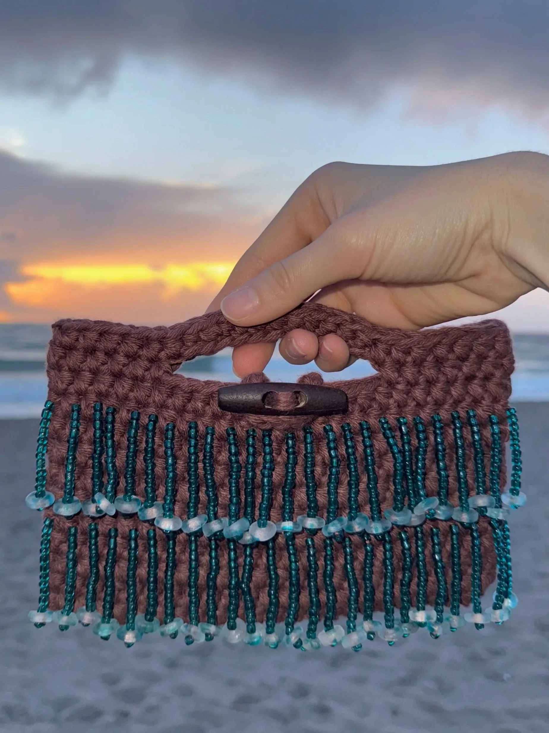Close-up of a hand holding a small woven handbag with beaded fringe details, against a backdrop of a beach at sunset with dark clouds and an orange and yellow sky.