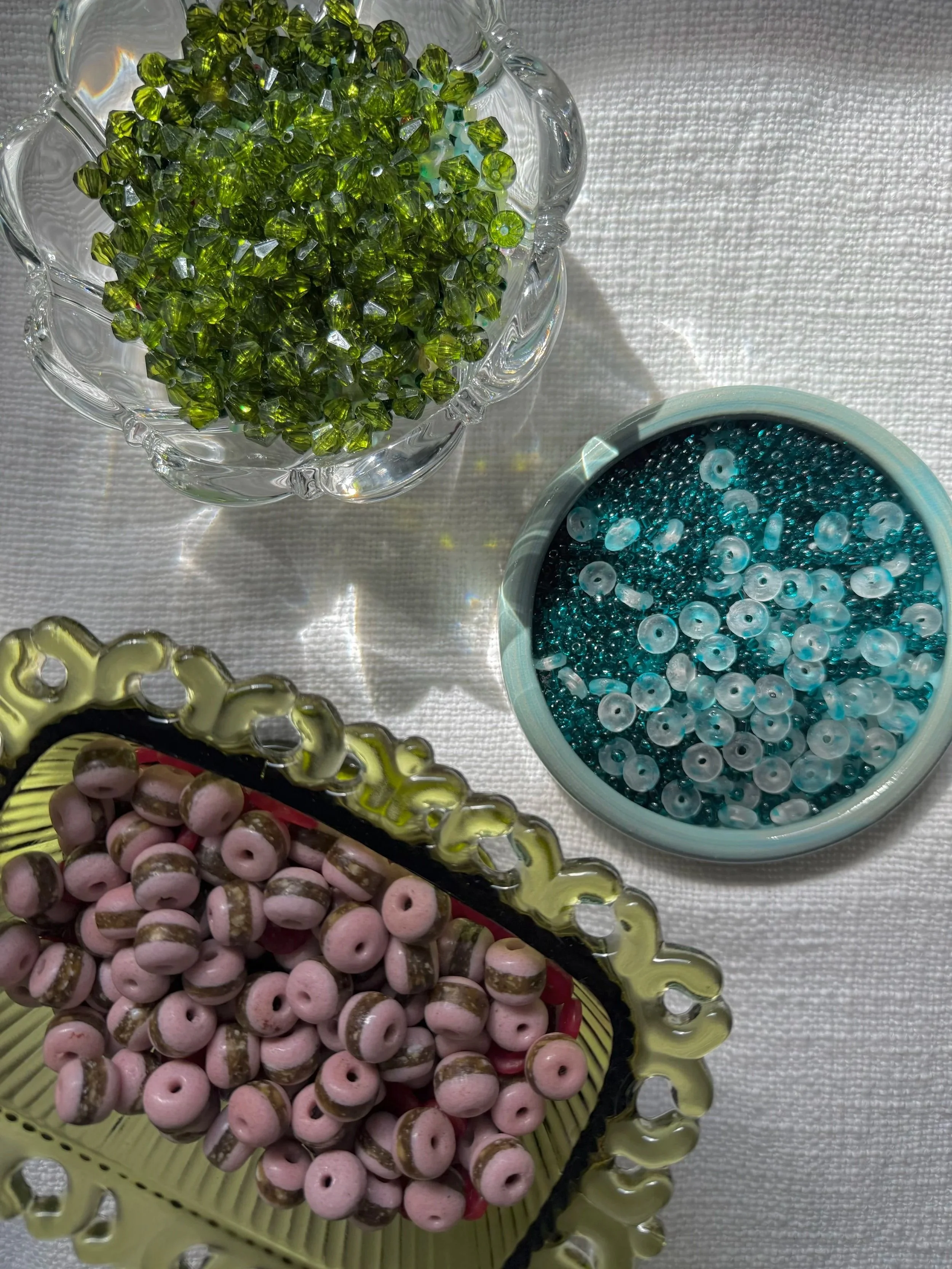 A collection of colorful beads in small bowls and a tray. The green beads are in a glass bowl, the blue beads are in a white container, and the pink beads are on a yellow tray with a decorative edge.