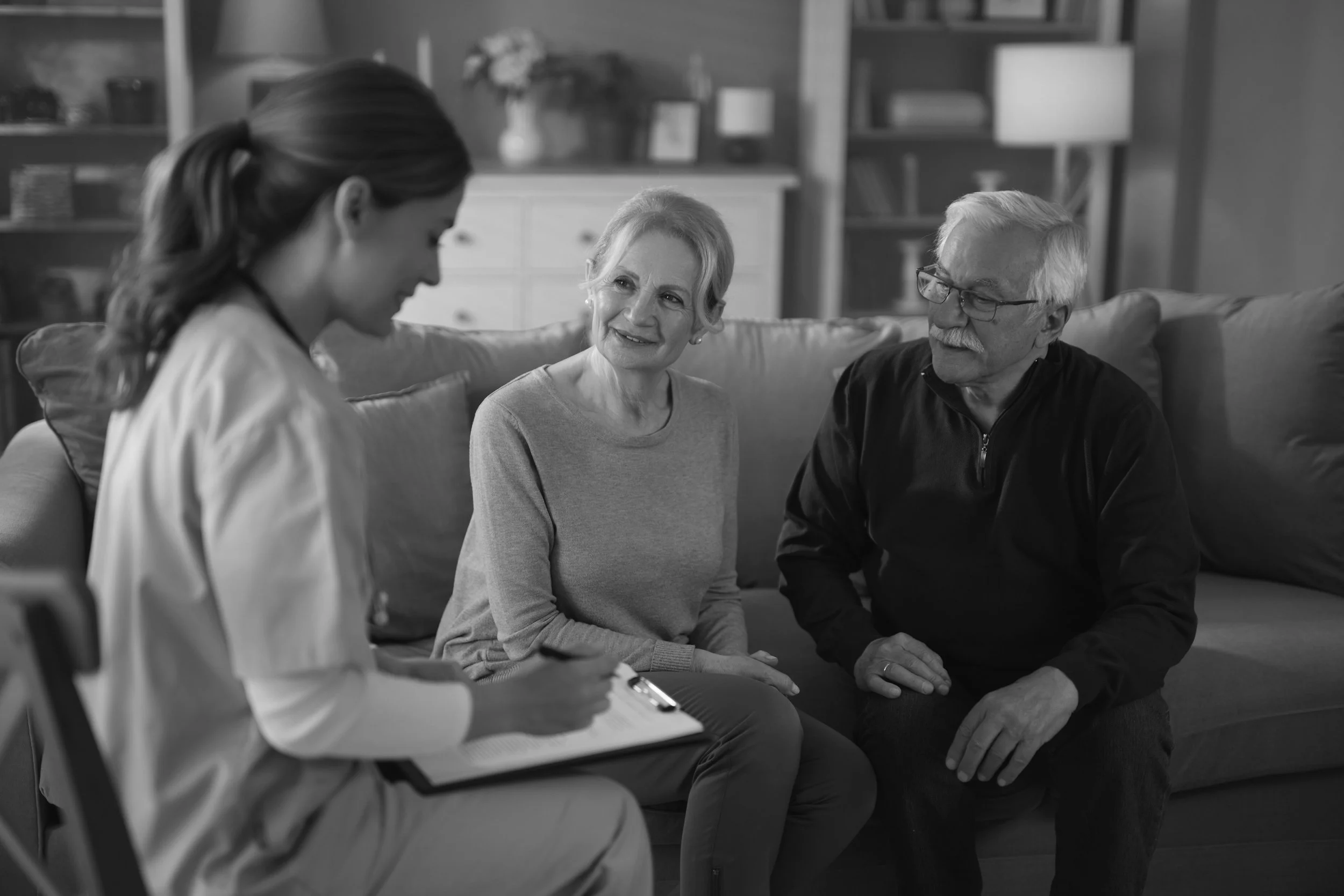 A woman and an elderly couple having a conversation in a living room, with the woman taking notes on a notepad.