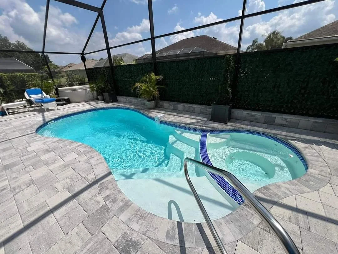Backyard swimming pool with a hot tub, surrounded by a stone deck, with lounge chairs and potted plants, enclosed by a screened enclosure under a partly cloudy sky.