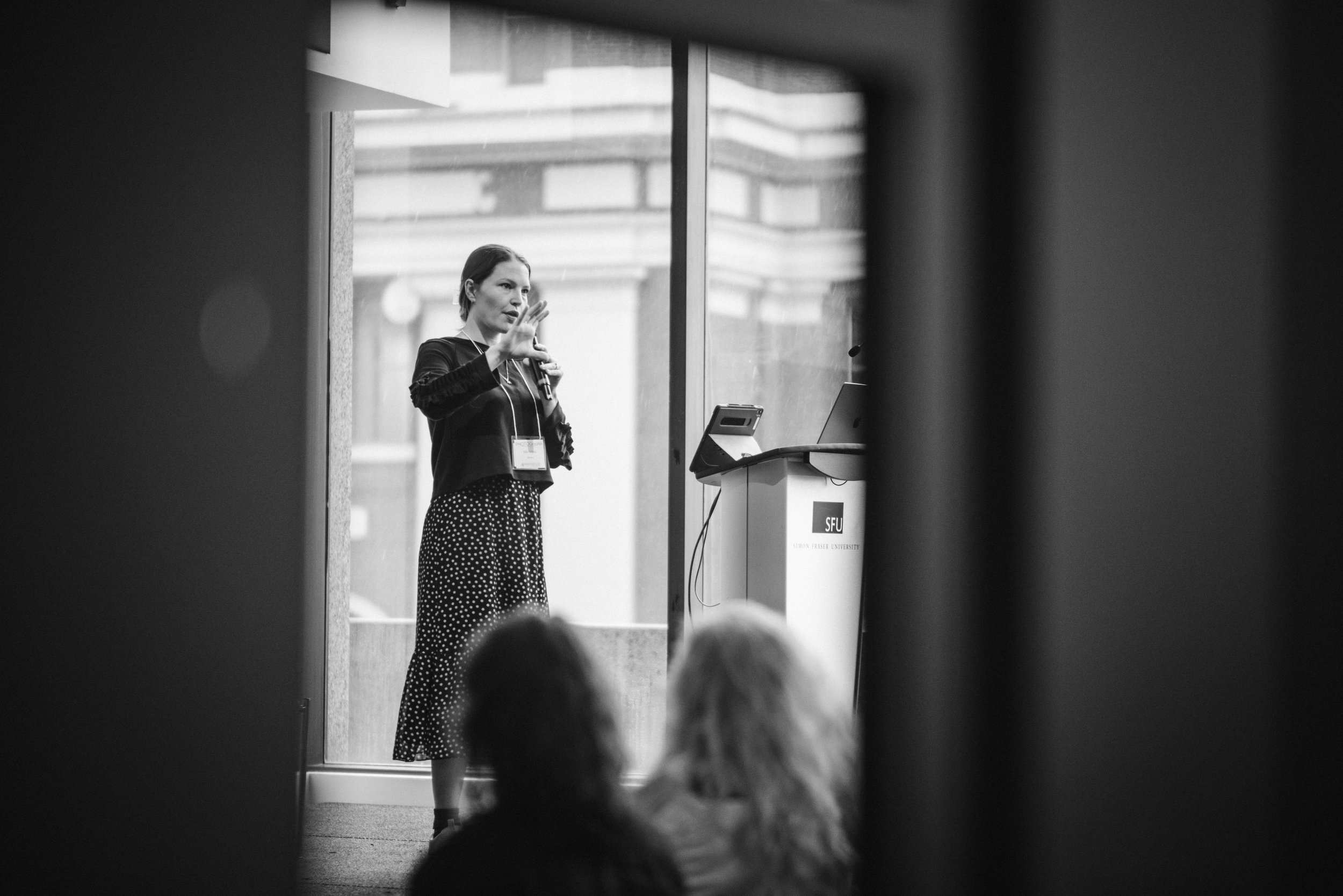Black and white photo of a woman speaking at a podium during a conference, viewed through a small opening or gap. The woman is wearing a polka dot skirt and a dark top, and is gesturing with one hand while holding a microphone with the other. The background shows a large window and a building outside.