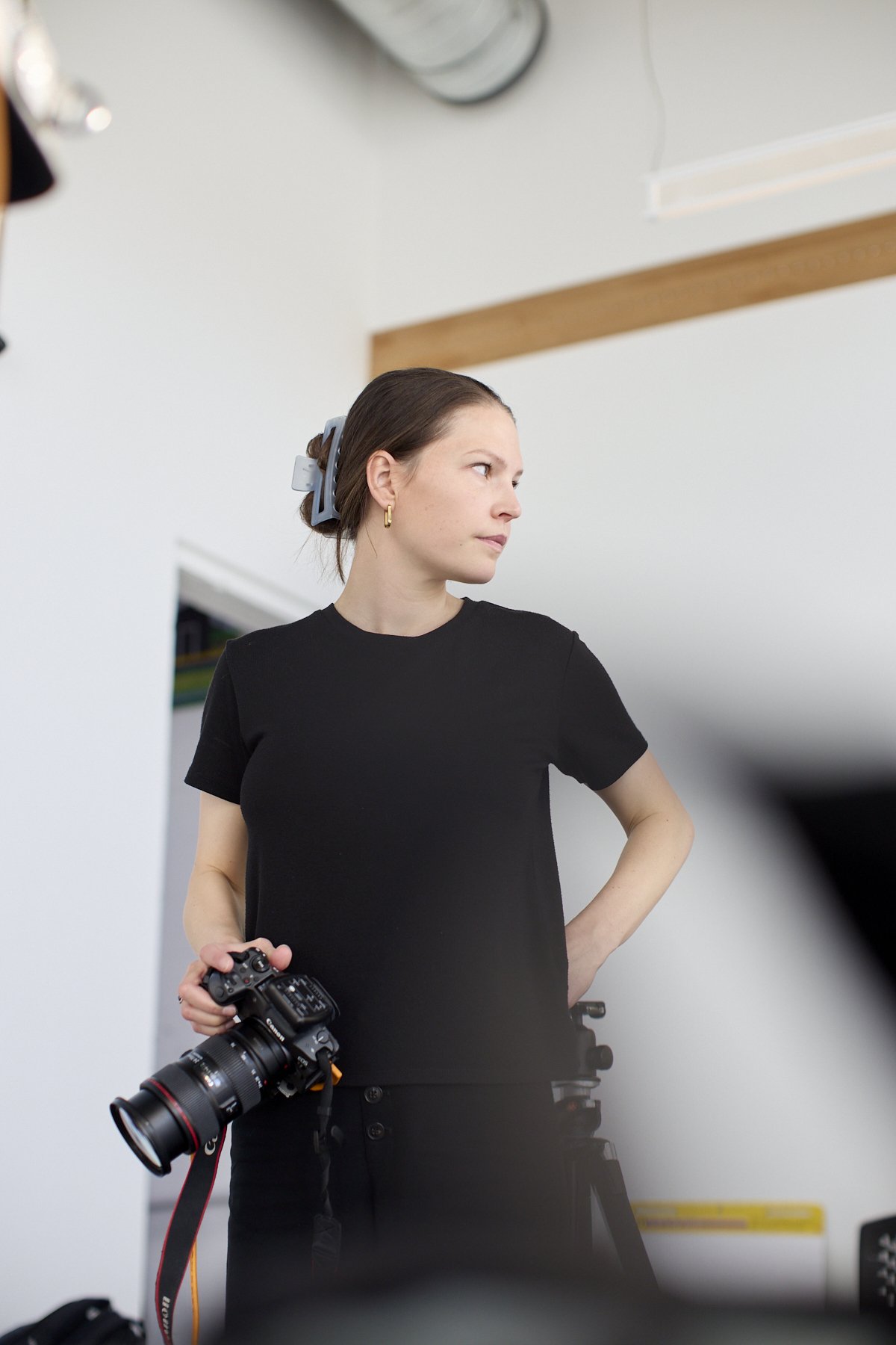 A young woman with brown hair tied back, wearing a black t-shirt and earrings, holds a professional camera with a large lens and a strap, standing indoors next to a tripod, with her left hand on her hip, looking to her right.