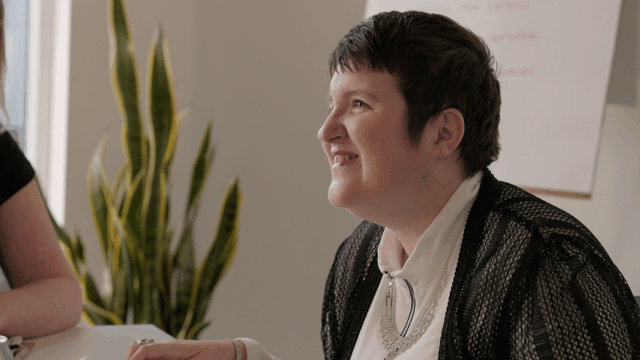 A person with short dark hair smiling, wearing a black and white jacket, sitting at a table in a room with a plant in the background.