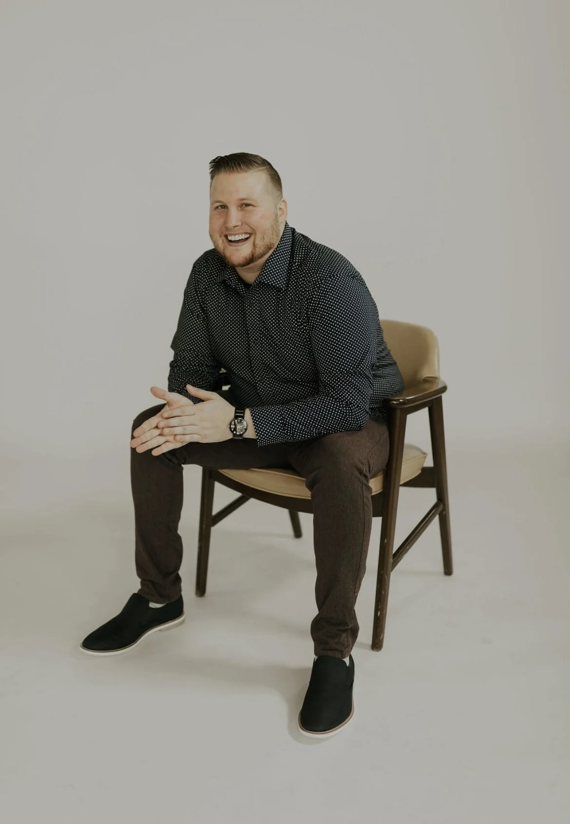 A man sitting on a wooden chair with a beige cushion, smiling, wearing a black polka dot shirt, brown pants, and black slip-on shoes, against a plain white background.