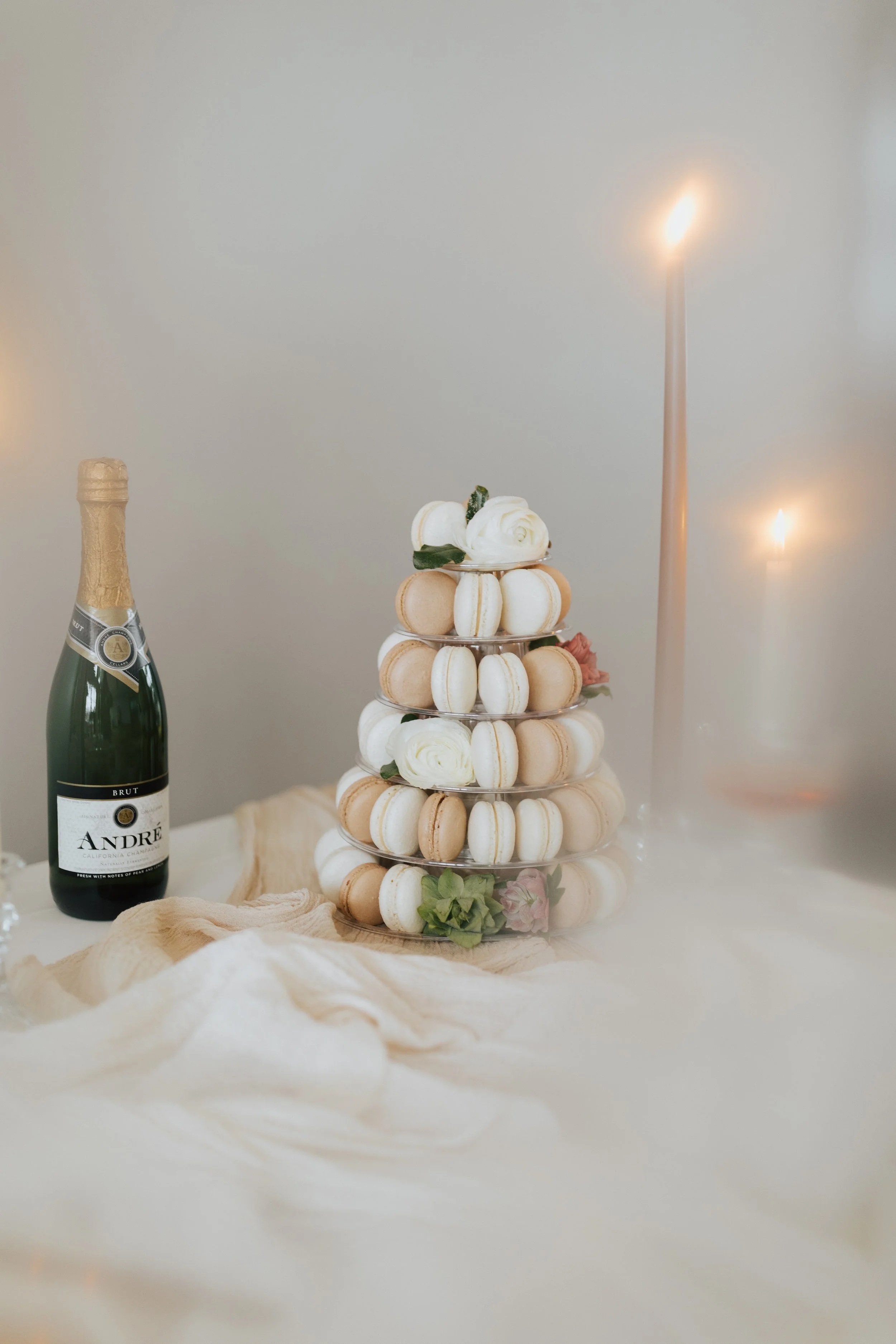 A champagne bottle and a layered display of macarons and flowers on a table with candles and soft fabric.