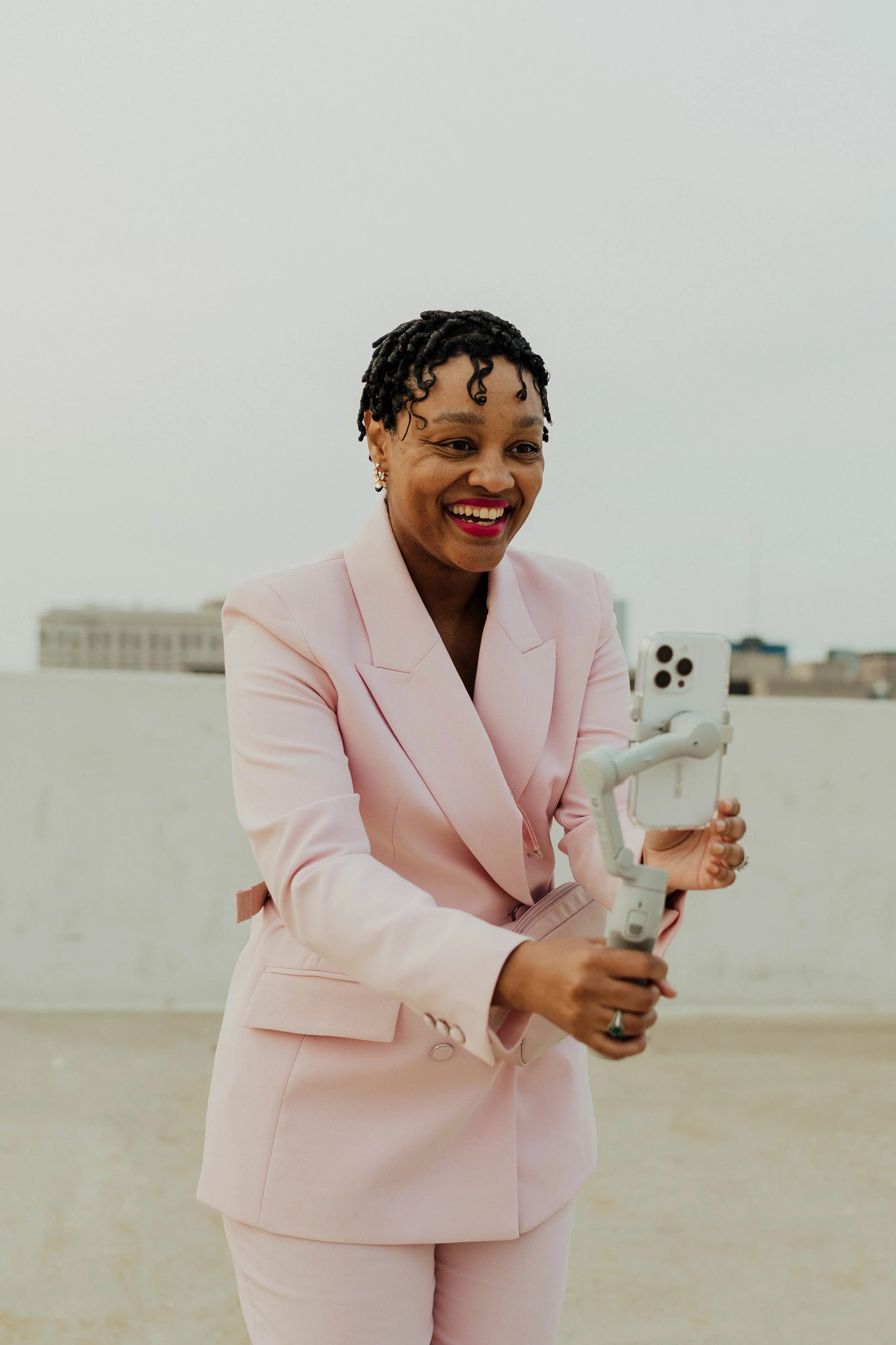 Woman in a pink suit smiling and recording herself with a smartphone on a stabilizing gimbal on a rooftop