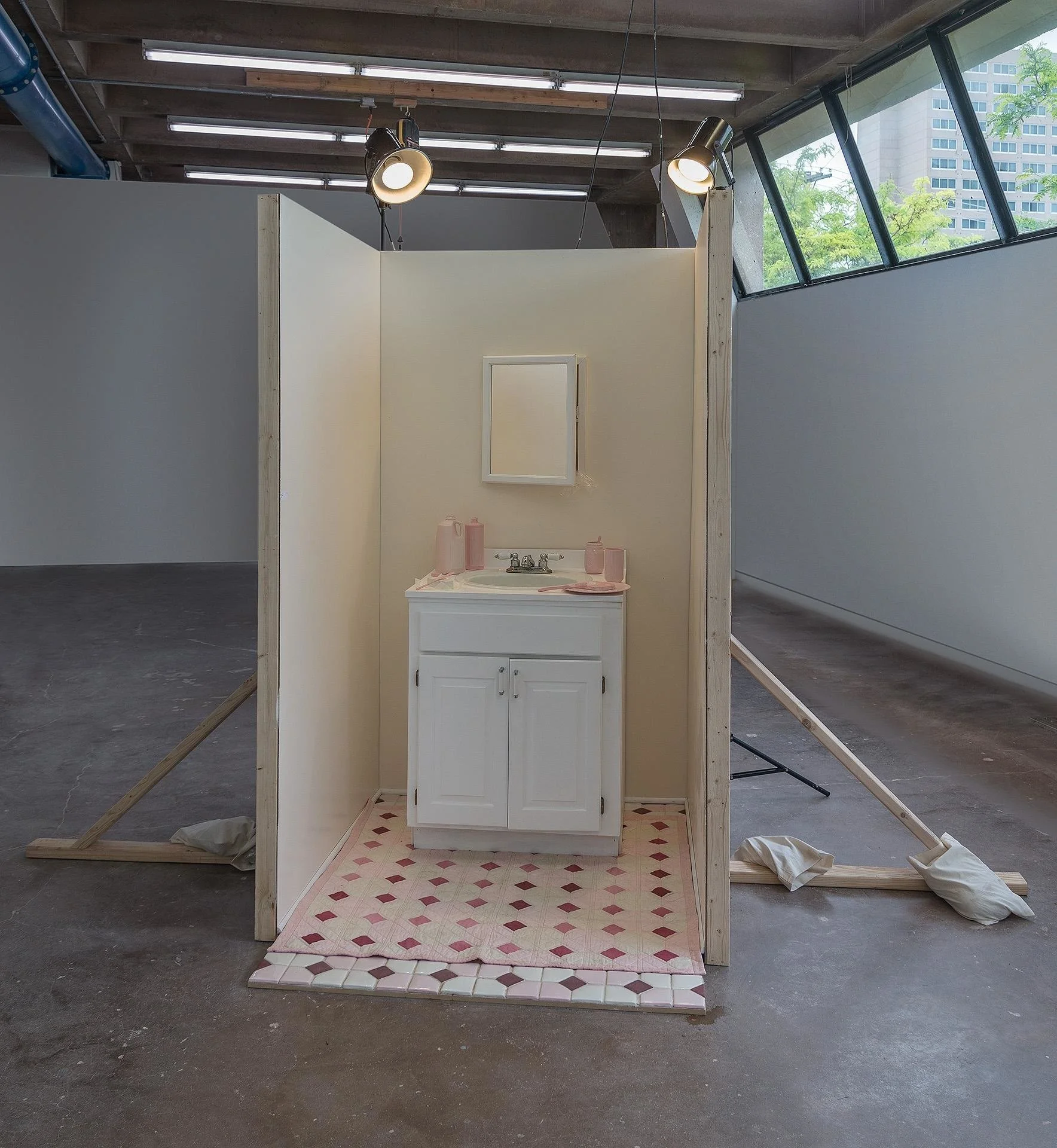 A front-view of "body map," a small bathroom built like a film set. The walls are a peachy color, and the white bathroom counter is littered with pink ceramic dishes, butter, and bread. The floor is tiled and covered with a quilt that matches.