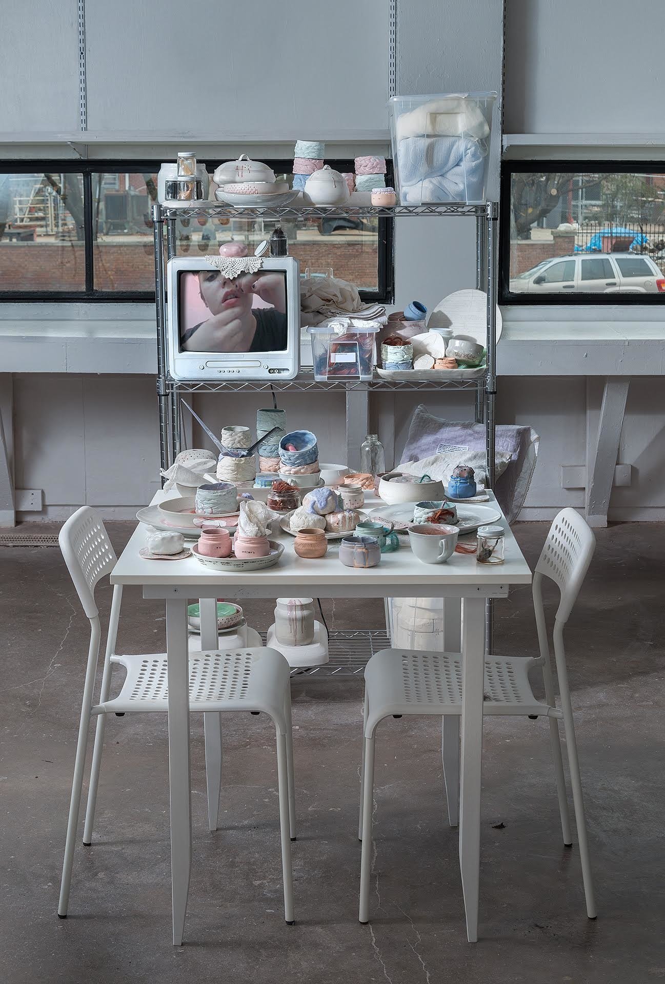 White table covered in wares in front of a metal shelf that is completely covered with stacked cups, urns that have pink glaze dripping off their lids, and a small box TV showing the artist threading hair through their labret.