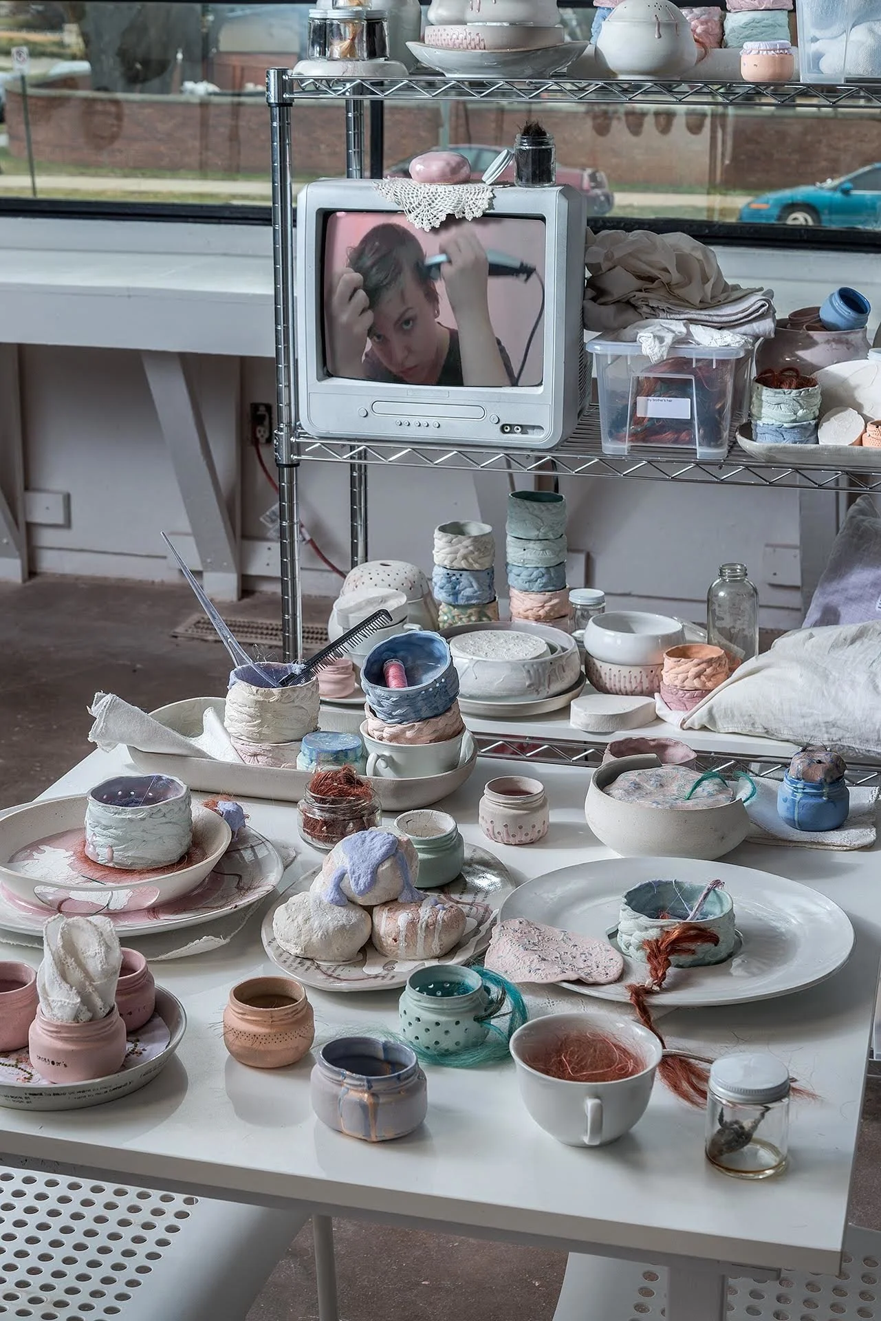 Detail with ceramic biscuits on an oval plate glazed with lines of text; slip-cast baby food jars in pink, orange, blue, and teal which are studded and pierced with short and long human hair; Ceramic embroidered napkins draped over dishes.